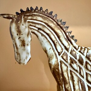 May include: A ceramic horse figurine with a cream-colored body and brown markings. The horse has a detailed mane and a textured pattern on its body. The figurine is a decorative item.