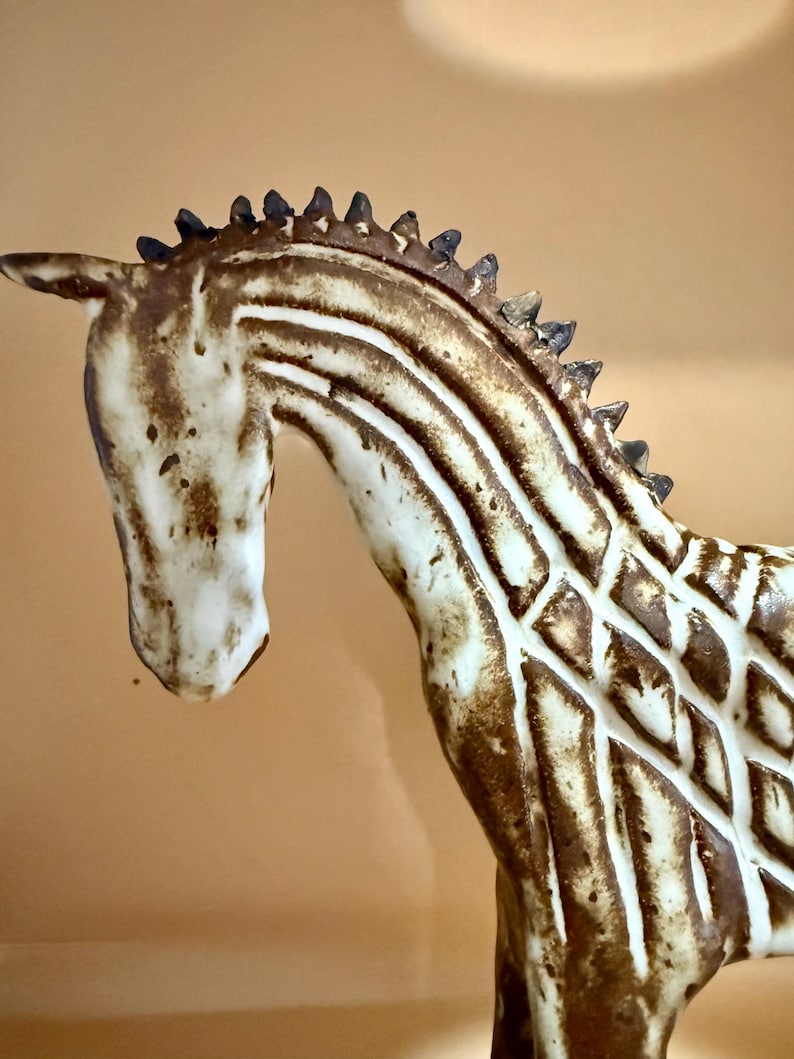 May include: A ceramic horse figurine with a cream-colored body and brown markings. The horse has a detailed mane and a textured pattern on its body. The figurine is a decorative item.
