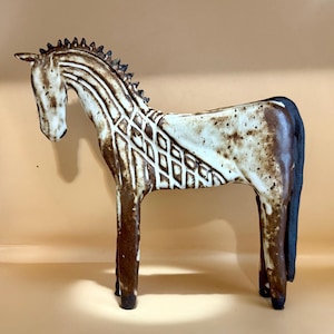 May include: A ceramic horse figurine in shades of brown and cream. The horse has a detailed mane and tail, with a geometric pattern on its body. The sculpture is standing, showcasing its elegant form and artistic design.