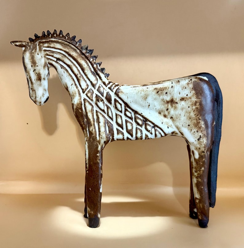 May include: A ceramic horse figurine in shades of brown and cream. The horse has a detailed mane and tail, with a geometric pattern on its body. The sculpture is standing, showcasing its elegant form and artistic design.