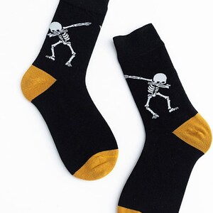 Puede incluir: Un par de calcetines negros con un dise&ntilde;o de esqueleto blanco haciendo un dab. Los talones y las puntas son dorados. Estos calcetines probablemente est&aacute;n hechos de un material suave y c&oacute;modo.