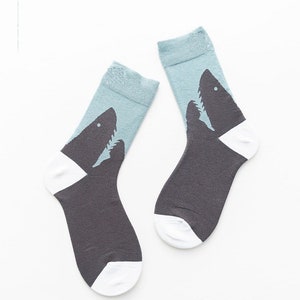 Puede incluir: Un par de calcetines con la silueta de un tiburón gris oscuro sobre un fondo azul claro. Los calcetines tienen talones y punteras blancas, con el diseño del tiburón extendiéndose desde el mediopié hasta el puño acanalado del calcetín.