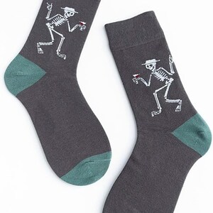 Puede incluir: Un par de calcetines gris oscuro con tal&oacute;n y puntera verde azulado. Cada calcet&iacute;n presenta un gr&aacute;fico de esqueleto blanco con sombrero y bebida. Los calcetines est&aacute;n hechos de un material suave.