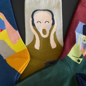 Puede incluir: Tres pares de calcetines con diferentes estampados artísticos. El primer par presenta un fondo azul con una figura amarilla y rosa. El segundo par tiene un fondo beige con una figura verde y rosa. El tercer par tiene un fondo rojo con una figura verde y negra.