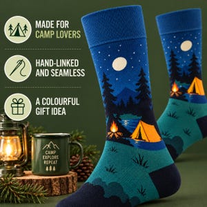 Puede incluir: Un par de calcetines azules con una escena de acampada nocturna que incluye una tienda de campaña amarilla, una hoguera, pinos, una luna y estrellas. Los calcetines tienen un puño azul oscuro y una base verde azulado. El texto dice: MADE FOR CAMP LOVERS, HAND-LINKED AND SEAMLESS, A COLOURFUL GIFT IDEA.