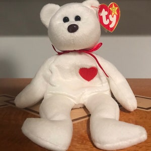 Puede incluir: Un oso de peluche blanco con un corazón rojo en el pecho y una cinta roja alrededor del cuello. El oso tiene ojos negros y una nariz marrón. Una etiqueta roja en forma de corazón está unida a su oreja.