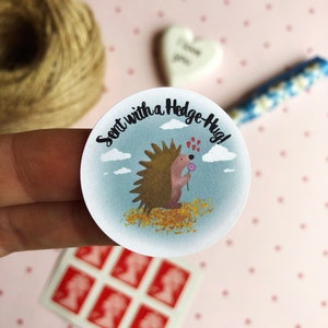 May include: A round sticker with a cartoon hedgehog holding a flower, surrounded by a blue sky and clouds. The text "Sent with a Hedge-Hug!" is written around the edge. The sticker is held by a hand, with stamps and twine in the background.