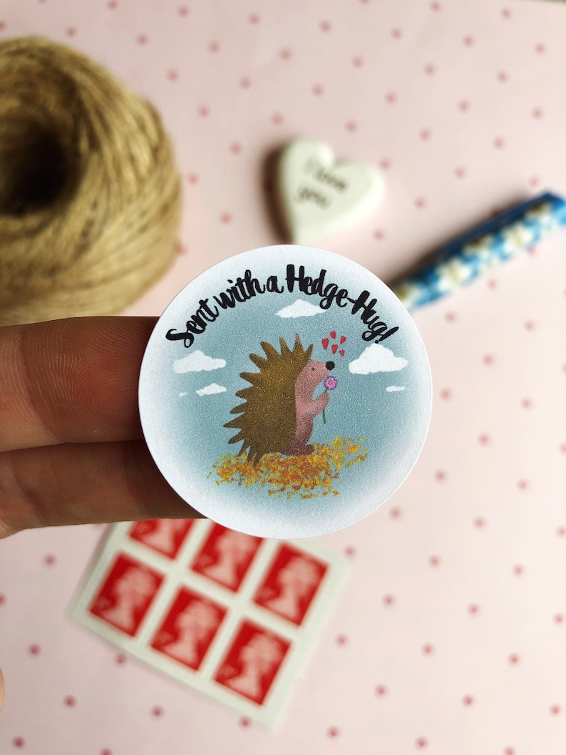 May include: A round sticker with a cartoon hedgehog holding a flower, surrounded by a blue sky and clouds. The text "Sent with a Hedge-Hug!" is written around the edge. The sticker is held by a hand, with stamps and twine in the background.