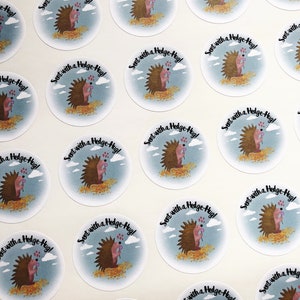 May include: Round stickers featuring a cartoon hedgehog with a pink nose and a sprig of flowers. The hedgehog is surrounded by a blue sky with clouds and text that reads "Sent with a Hedge-Hug!".