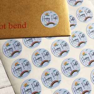 以下が含まれることがあります： 虹、太陽、雲、そして「Happy Post！」の文字が描かれた、水色の背景の丸いステッカーのシート。同じステッカーが付いた茶色の封筒が見えます。封筒には「not bend」の文字が印刷されています。
