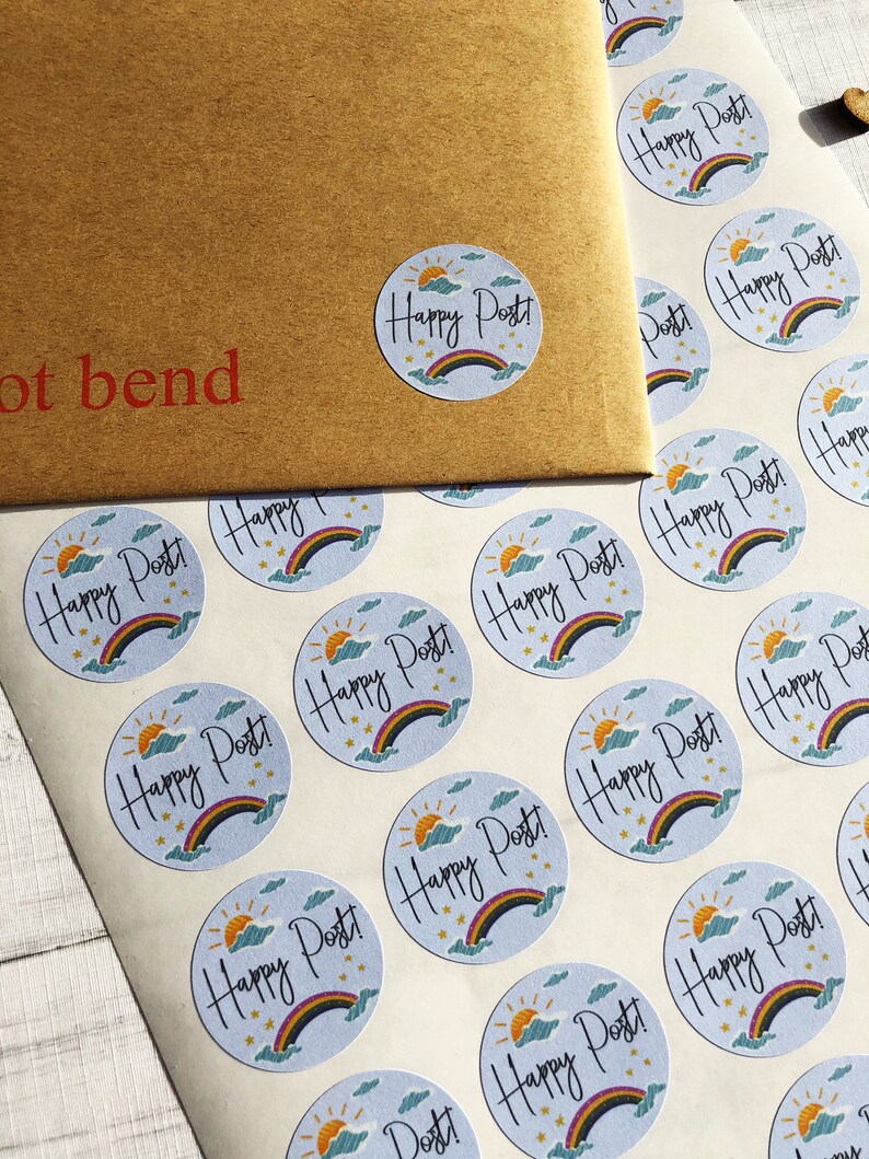 以下が含まれることがあります： 虹、太陽、雲、そして「Happy Post！」の文字が描かれた、水色の背景の丸いステッカーのシート。同じステッカーが付いた茶色の封筒が見えます。封筒には「not bend」の文字が印刷されています。