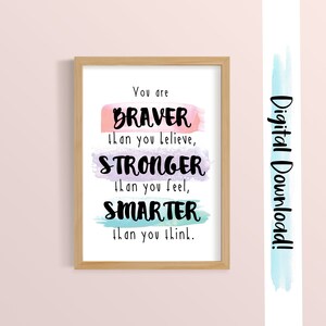 May include: A printable inspirational quote in a wooden frame. The quote reads "You are BRAVER than you believe, STRONGER than you feel, SMARTER than you think." The words are written in black and are layered over watercolor washes in pink, purple, and blue.