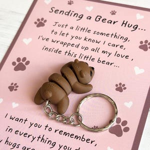 May include: A brown bear-shaped keyring with a silver key ring and a pink card with the text "Sending a Bear Hug... Just a little something, to let you know I care, I've wrapped up all my love, inside this little bear... I want you to remember, in everything you hugs are"