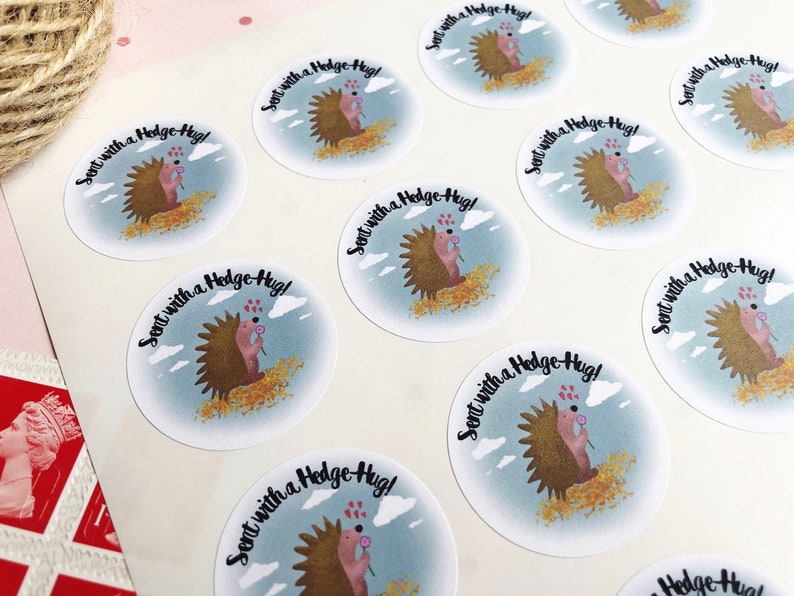 May include: Round stickers with a cartoon hedgehog design. The hedgehog is brown with a pink nose and is holding a flower. The stickers have the text "Sent with a Hedge-Hug!" on a light blue background with clouds.
