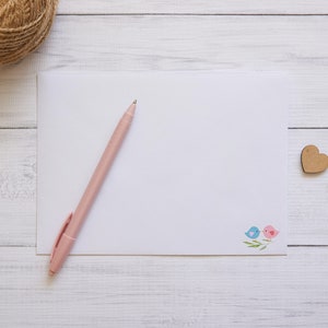 May include: A blank white card with a pink pen on top of it. The card has a small design of two birds on a branch in the bottom right corner. A wooden heart is on the right side of the card. The card and pen are on a white wooden surface.