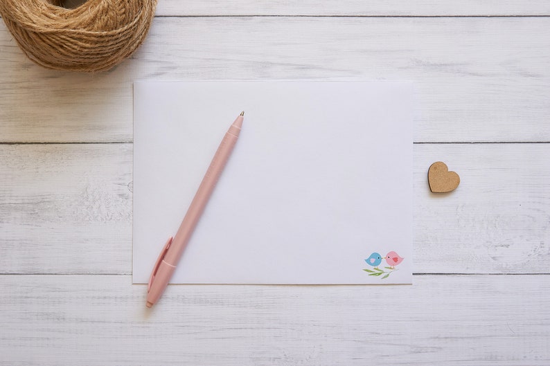 May include: A blank white card with a pink pen on top of it. The card has a small design of two birds on a branch in the bottom right corner. A wooden heart is on the right side of the card. The card and pen are on a white wooden surface.