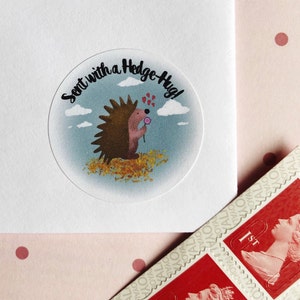 May include: A clear, circular sticker with a cartoon hedgehog holding a flower, surrounded by the text "Sent with a Hedge-Hug!". The sticker is on a white envelope, with red stamps in the corner. The background is a light pink with red polka dots.