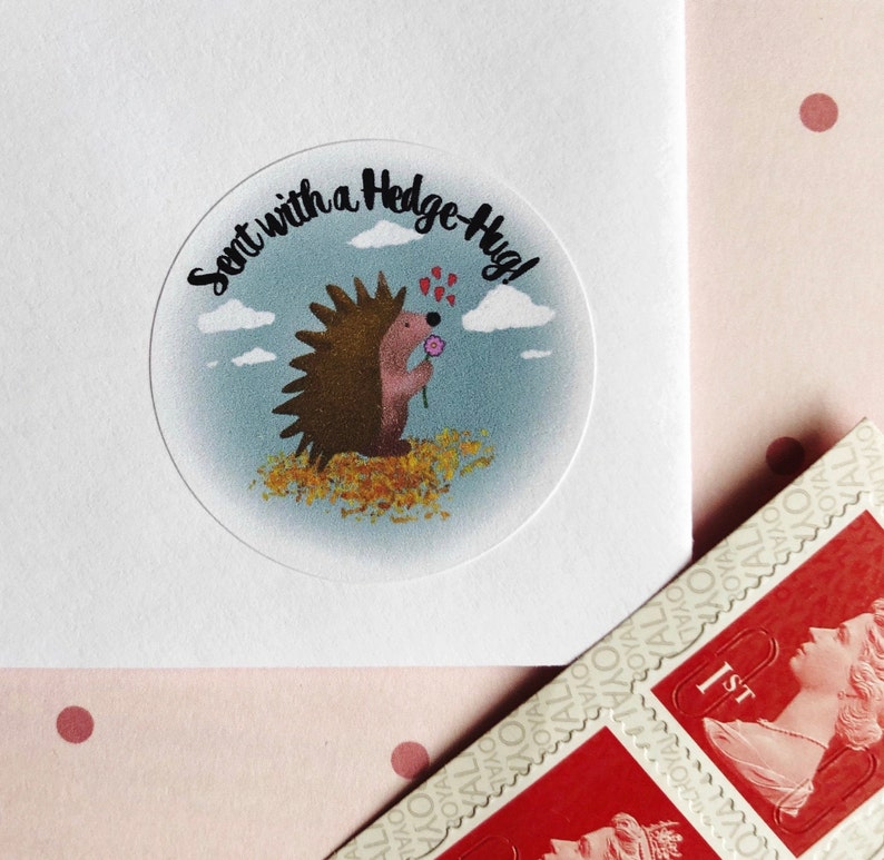 May include: A clear, circular sticker with a cartoon hedgehog holding a flower, surrounded by the text "Sent with a Hedge-Hug!". The sticker is on a white envelope, with red stamps in the corner. The background is a light pink with red polka dots.