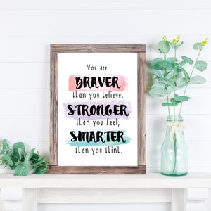 May include: A framed print with a motivational quote: "You are BRAVER than you believe, STRONGER than you feel, SMARTER than you think." The quote is written in black on a background of watercolor washes in pink, purple, and blue.