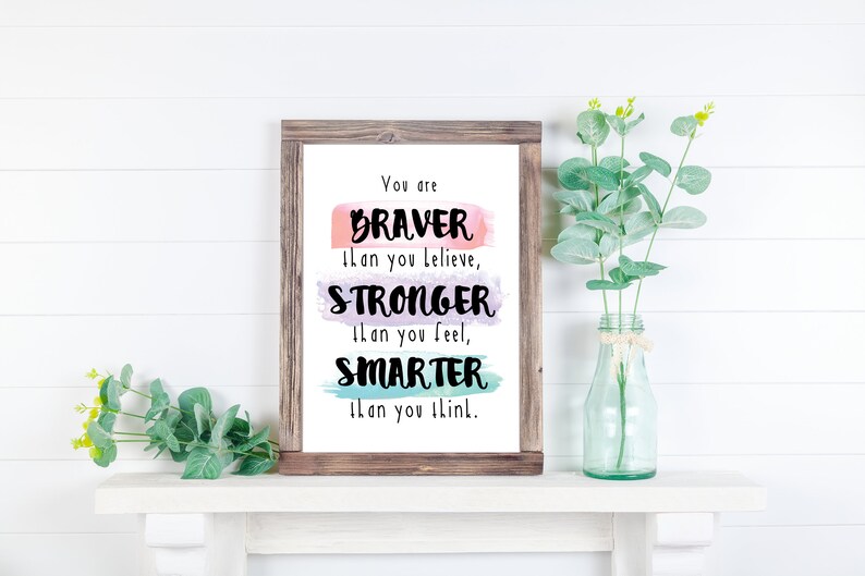 May include: A framed print with a motivational quote: "You are BRAVER than you believe, STRONGER than you feel, SMARTER than you think." The quote is written in black on a background of watercolor washes in pink, purple, and blue.