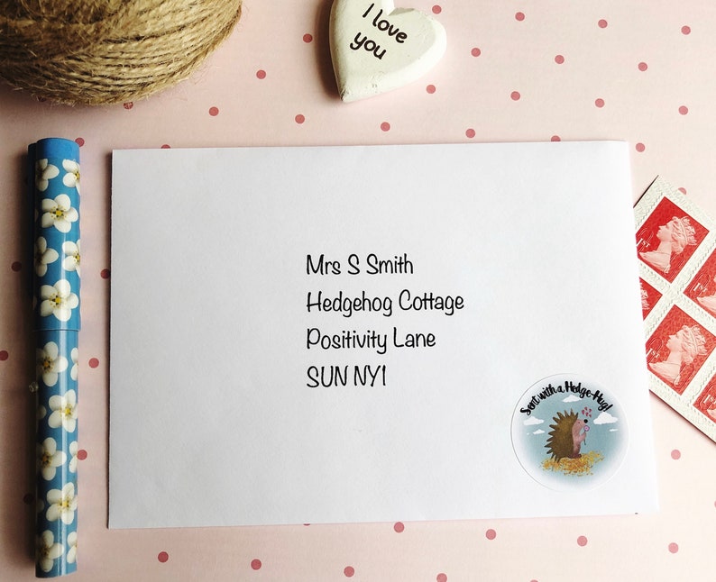 May include: A white envelope with an address label, a blue pen with white flowers, and a decorative heart that says "I love you". The envelope has the address "Mrs S Smith, Hedgehog Cottage, Positivity Lane, SUN NYI". A sticker says "Sent with a Hedge Hug!"