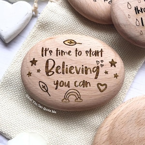 May include: A smooth, oval-shaped wooden stone featuring laser-engraved text that reads, "It's time to start Believing you can." The design includes small engraved icons of stars, leaves, a heart, and a rainbow, with the text "And You Can Quote Me" visible at the bottom left.
