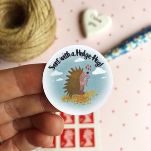 May include: A round sticker with a cartoon hedgehog holding a flower, surrounded by the text "Sent with a Hedge-Hug!" The hedgehog is brown with a pink face and is set against a blue sky background. The sticker is held in a hand.