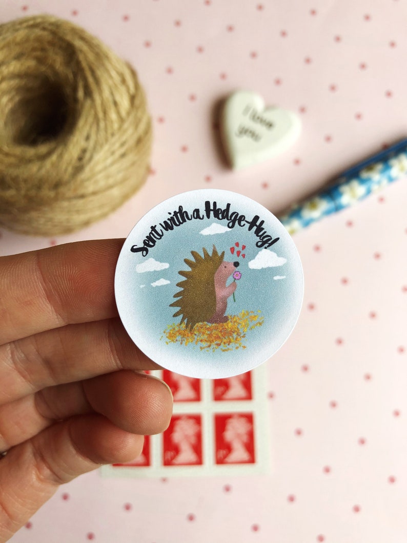 May include: A round sticker with a cartoon hedgehog holding a flower, surrounded by the text "Sent with a Hedge-Hug!" The hedgehog is brown with a pink face and is set against a blue sky background. The sticker is held in a hand.