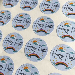 以下が含まれることがあります： 水色の背景に「Happy Post!」の文字が筆記体で書かれた丸いステッカーのシート。虹、太陽、雲、星のイラストも描かれています。