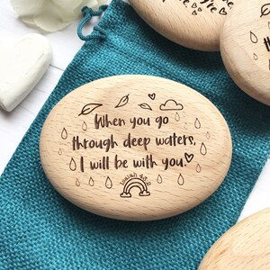 Puede incluir: Ficha de madera ovalada con el texto "When you go through deep waters, I will be with you." y un arco&iacute;ris con el texto "Isaiah 43:2" debajo. La ficha est&aacute; rodeada de peque&ntilde;as gotas de lluvia y hojas.