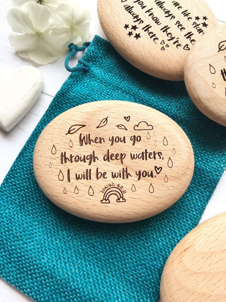Puede incluir: Ficha de madera ovalada con el texto "When you go through deep waters, I will be with you." y un arco&iacute;ris con el texto "Isaiah 43:2" debajo. La ficha est&aacute; rodeada de peque&ntilde;as gotas de lluvia y hojas.