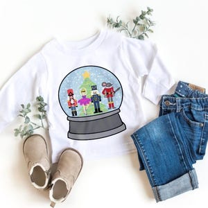 Może przedstawiać: Biała koszulka z długim rękawem z motywem kuli śnieżnej z choinką i żołnierzykami. Obraz zawiera również niebieskie dżinsy i beżowe buciki dla dziecka, tworząc świąteczny motyw.