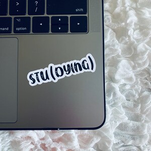 May include: A silver laptop computer with a white sticker that says "STU (DYING)" on the top right corner.