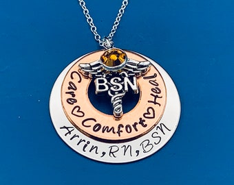 May include: Silver and copper necklace with a medical caduceus charm, a yellow rhinestone, and the words "Care Comfort Heal" and "Arrin, RN, BSN".