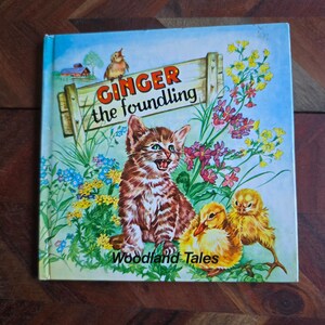 May include: A vintage children's book cover with a blue background and a sign that reads "Ginger the foundling". The cover features a brown and white tabby kitten, two yellow chicks, and a variety of colorful flowers. The book title is "Woodland Tales".