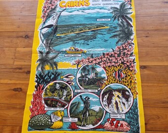 Op de afbeelding: Een gele theedoek met een kleurrijke illustratie van Cairns, Australië. De illustratie bevat een marlijn, Green Island, Stoney Creek Falls, het standbeeld van Captain Cook, The Esplanade en Barron Falls. De tekst "Cairns, Great Barrier Reef, North Queensland" staat bovenaan de theedoek gedrukt.