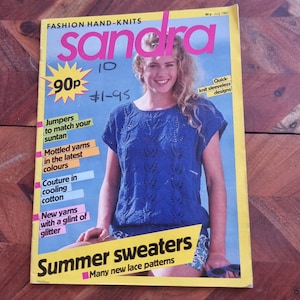 Op de afbeelding: Een tijdschriftcover voor Sandra, een tijdschrift voor handgebreide mode, met een model dat een blauwe gebreide zomertrui draagt met een kantpatroon. De tekst op de omslag luidt "Zomertruien" en "Veel nieuwe kantpatronen".