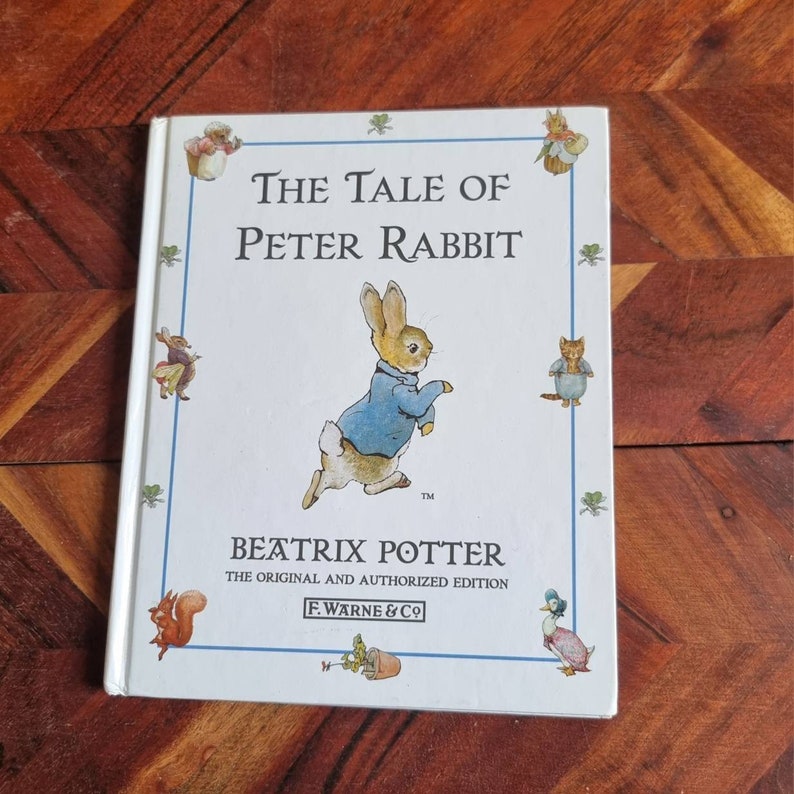 May include: A white book cover featuring a blue and white illustration of Peter Rabbit running. The title "The Tale of Peter Rabbit" is printed in black text. The author, Beatrix Potter, is printed below the illustration. The book is the original and authorised edition published by F. Warne & Co.