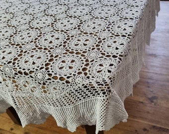Könnte beinhalten: Weiße Häkeltischdecke mit einem zarten Blumenmuster. Die Tischdecke hat einen gewellten Rand und ist über einen Holztisch gelegt.