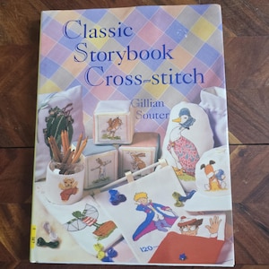 May include: A book titled "Classic Storybook Cross-stitch" with a cover showing cross-stitch examples. The examples include a duck, a bear, and a boy in a blue coat. The book is surrounded by cross-stitch projects and pencils.