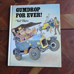 May include: A vintage children's book titled "Gumdrop Forever!" by Val Biro. The cover features a colorful illustration of a blue vintage car with passengers, including a dog, and a yellow car in the background. The book is on a wooden surface.