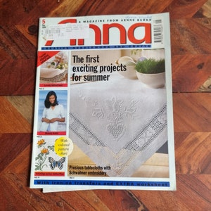 Op de afbeelding: Een tijdschriftcover voor "Anna, Creatief Naaldwerk en Handwerk" met een wit tafelkleed met delicate borduurwerk. De tekst op de omslag luidt "De eerste spannende projecten voor de zomer" en "Kostbare tafelkleden met Schwalmer borduurwerk".