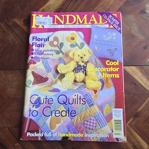 以下が含まれることがあります： 「HANDMADE」というタイトルの雑誌の表紙で、「CRAFT DECORATING SEWING」というサブタイトルが付いています。パッチワークのキルトの上にテディベアがいます。「Floral Flair」と「Cute Quilts to Create」というテキストと「132-PAGE ISSUE」のバナーが含まれています。