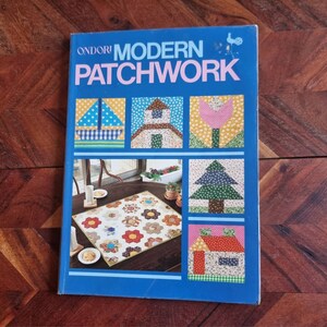 Op de afbeelding: Een blauw boek getiteld "ONDORI MODERN PATCHWORK" met kleurrijke patchwork-ontwerpen op de omslag. De ontwerpen omvatten een zeilboot, een huis, een tulp, een boom en een ander huis.
