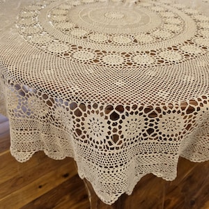 Könnte beinhalten: Eine runde, cremefarbene Häkel-Tischdecke mit einem komplizierten, sich wiederholenden Blumenmuster in der Mitte, das in ein feines Netzgitter übergeht, abgeschlossen mit einem gewellten Rand und einer breiten, detaillierten Bordüre.
