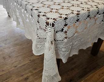 Könnte beinhalten: Weiße Häkeltischdecke mit einem zarten Blumenmuster. Die Tischdecke ist aus einem feinen, leichten Material gefertigt und hat einen gewellten Rand.