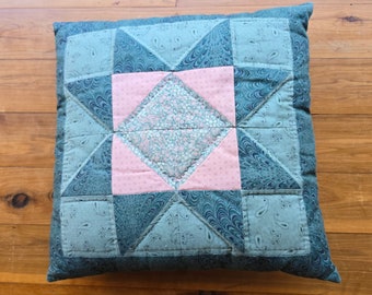 May include: A square pillow with a patchwork design. The pillow is made of different shades of blue and pink fabric. The center of the pillow features a large pink square with a smaller blue and pink square inside.