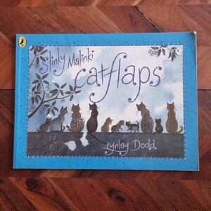 May include: A blue book cover with the title "Slinky Malinki Catflaps" in white cursive lettering. The book cover features a watercolor illustration of a group of cats with a tree branch in the background. The author's name, "Lynley Dodd", is printed in white below the illustration.
