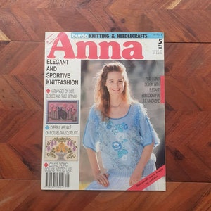 Op de afbeelding: Een tijdschriftcover voor Burda Breien & Handwerken Anna, met een vrouw die een lichtblauwe gebreide trui draagt met bloemenborduurwerk. De tekst op de cover luidt "Elegant en sportief breien" en bevat details over de ontwerpen in de binnenkant, zoals "Hardanger op rok, blouses en tafelkleden" en "Vrolijke applicaties op foto's, tafelkleden, etc."