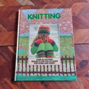 May include: A vintage knitting book with a green, brown, and white cover. The cover features a photo of a young girl wearing a knitted hat, mittens, and socks. The book title is "Knitting" and the text below the photo reads "Over 20 exciting projects for you to make for home and family."